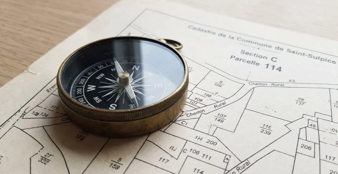 Gros plan sur une carte cadastrale d'un domaine avec une boussole vintage posée dessus, lumière naturelle rasante créant des ombres douces